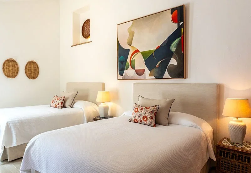 A bedroom with two beds, each with decorative pillows, nightstands with lamps, and abstract artwork on the wall.