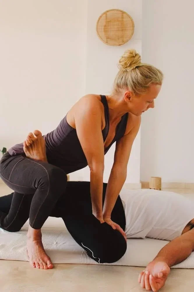A woman practicing yoga in a yoga studio or home, with a person lying on the floor next to her, both on a yoga mat.