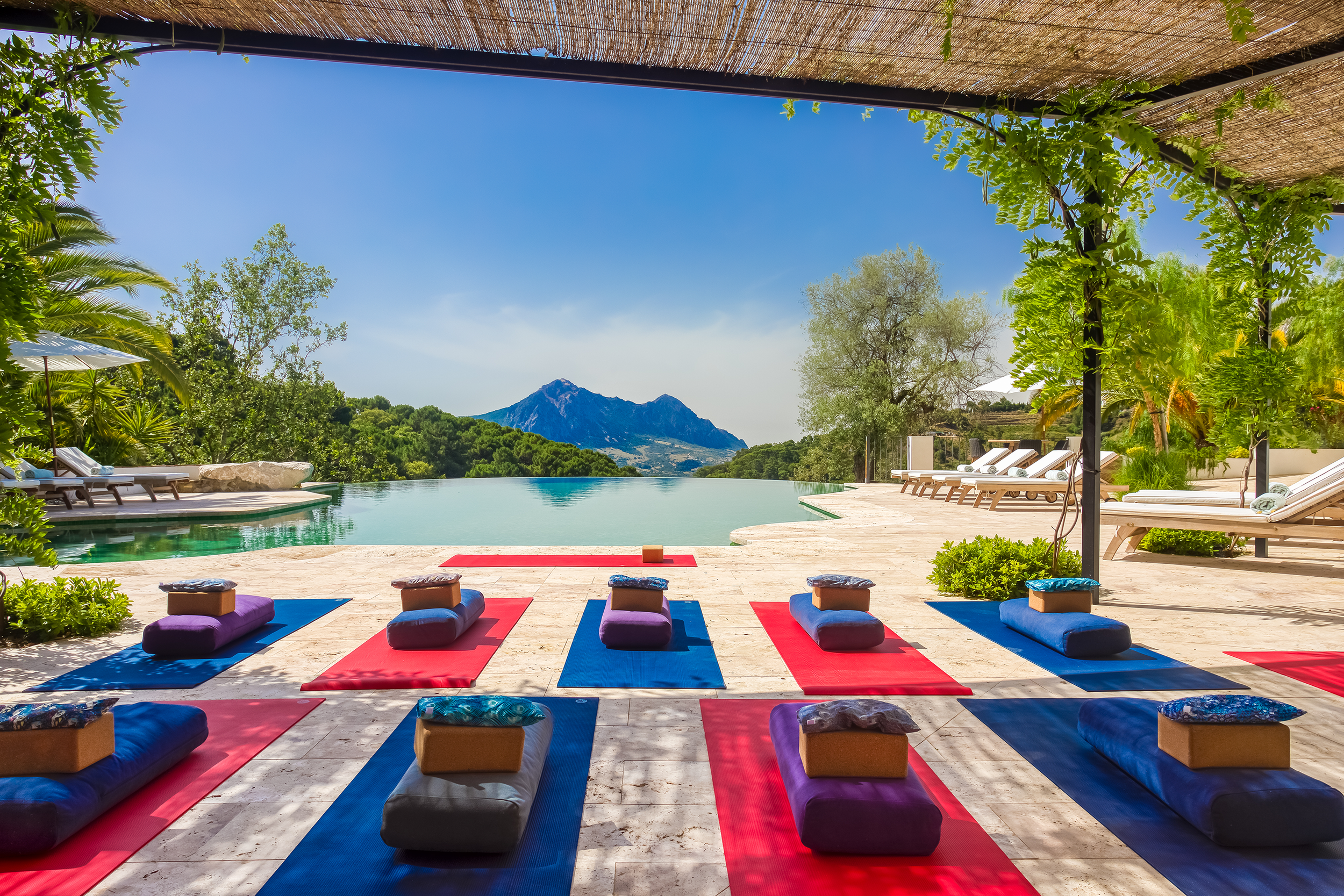 Yoga mats and cushions arranged on a deck overlooking an infinity pool with a mountain and trees in the background, shaded by a thatched roof.
