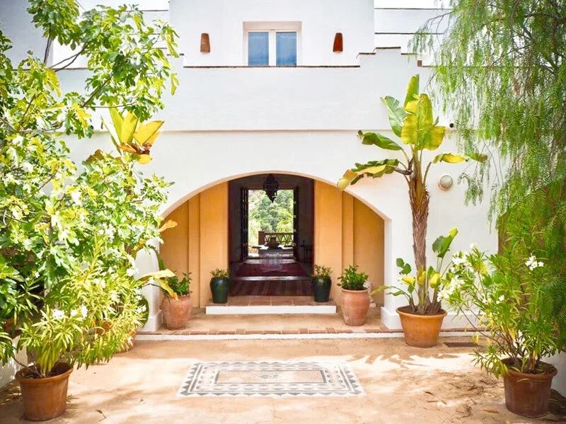 Front entrance of a house with an archway, surrounded by potted plants, with a view toward an outdoor seating area in the background.