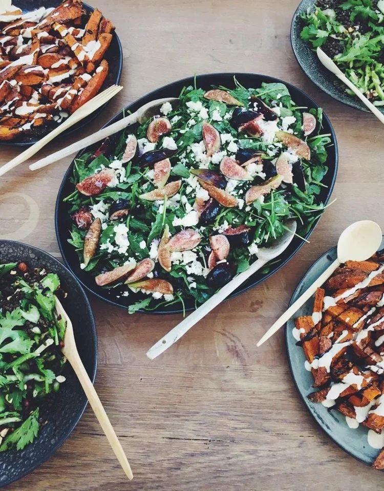 Salad with figs, olives, feta cheese, and greens on a black platter, and plates with grilled meat and waffle fries drizzled with sauce on a wooden table.