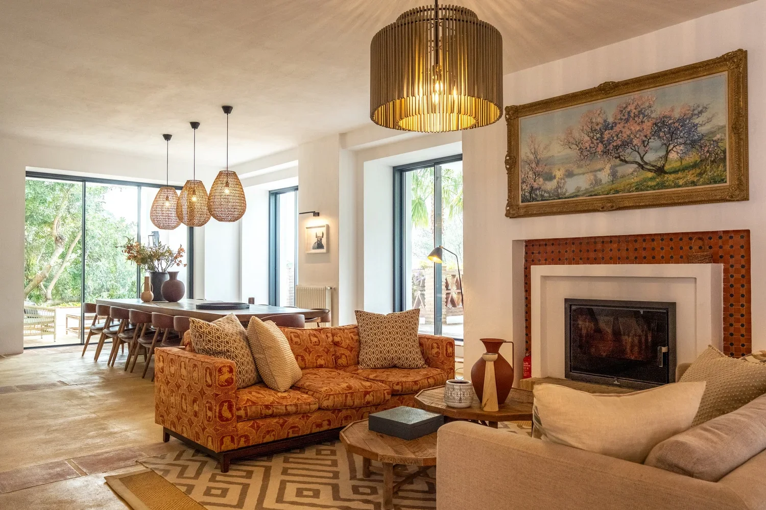 Interior of a living and dining area with large windows, a patterned sofa with pillows, a wooden coffee table, a fireplace with a painting above, and a dining table with seating, decorated with vases and pendant lights.
