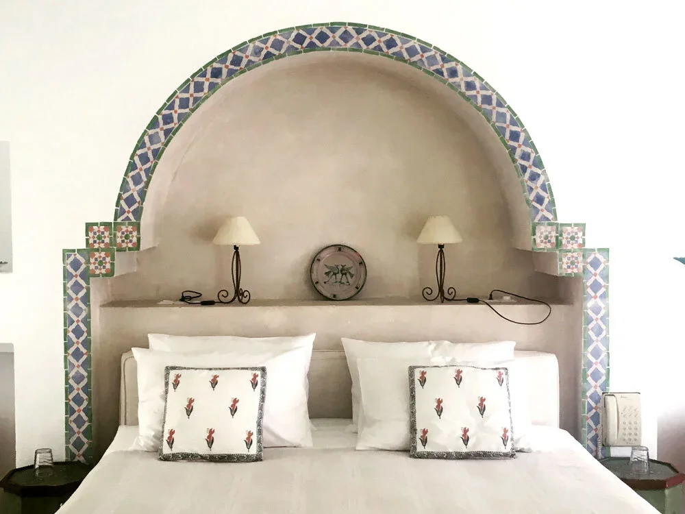 A bedroom with an arched decorative wall behind the bed, featuring colorful tile patterns. The bed has white linens and pillows with red flower motifs. Two wall-mounted lamps with beige shades are on either side of the clock on the shelf above the be