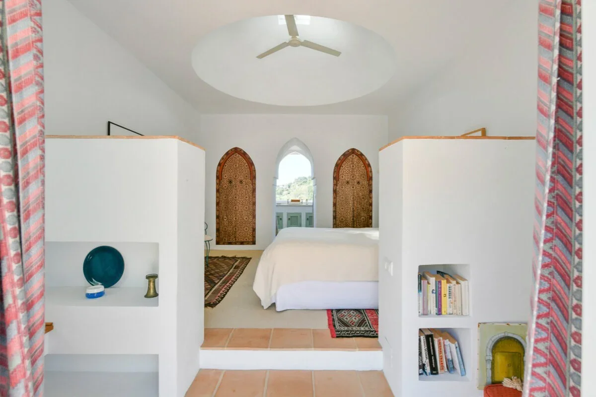 A cozy bedroom with white walls and a ceiling fan, a bed with white bedding, colorful rugs, and built-in shelves with books and decorative items, framed by red-pink patterned curtains.