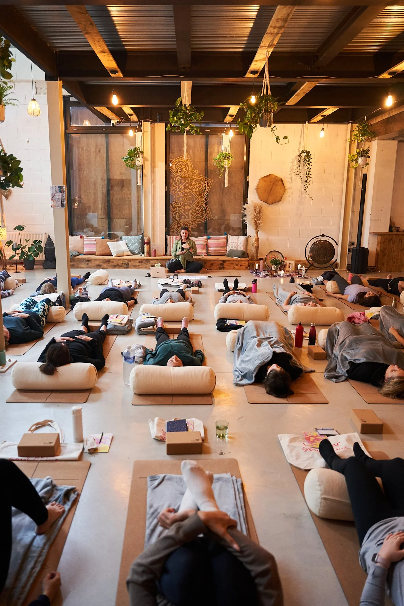 Participants lying on yoga mats with bolster pillows in a warmly lit, cozy yoga studio during a class led by an instructor sitting cross-legged at the front.