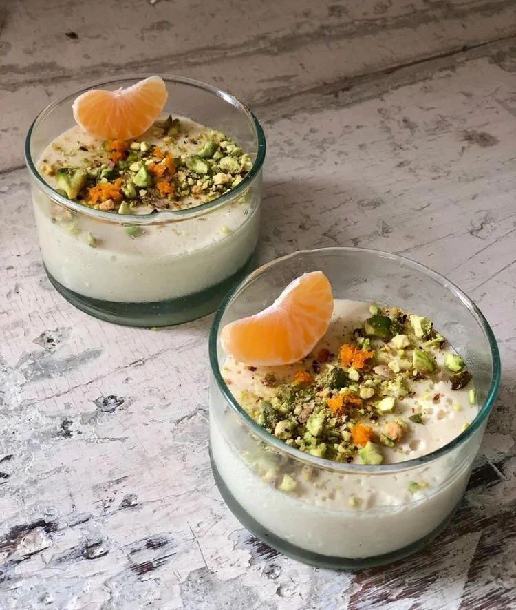 Two glasses of pistachio pudding topped with orange segments and chopped pistachios on a rustic wooden surface.