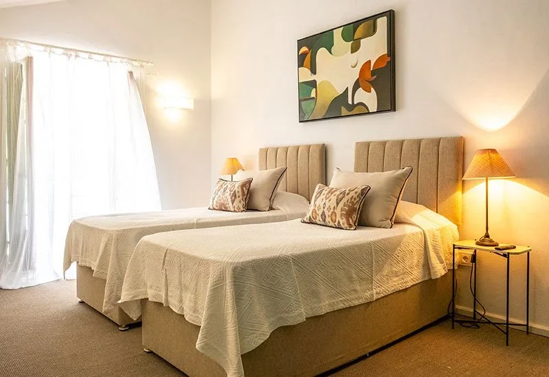 A hotel room with two beds, beige headboards, decorative pillows, a nightstand with a lamp, and a colorful abstract painting on the wall.