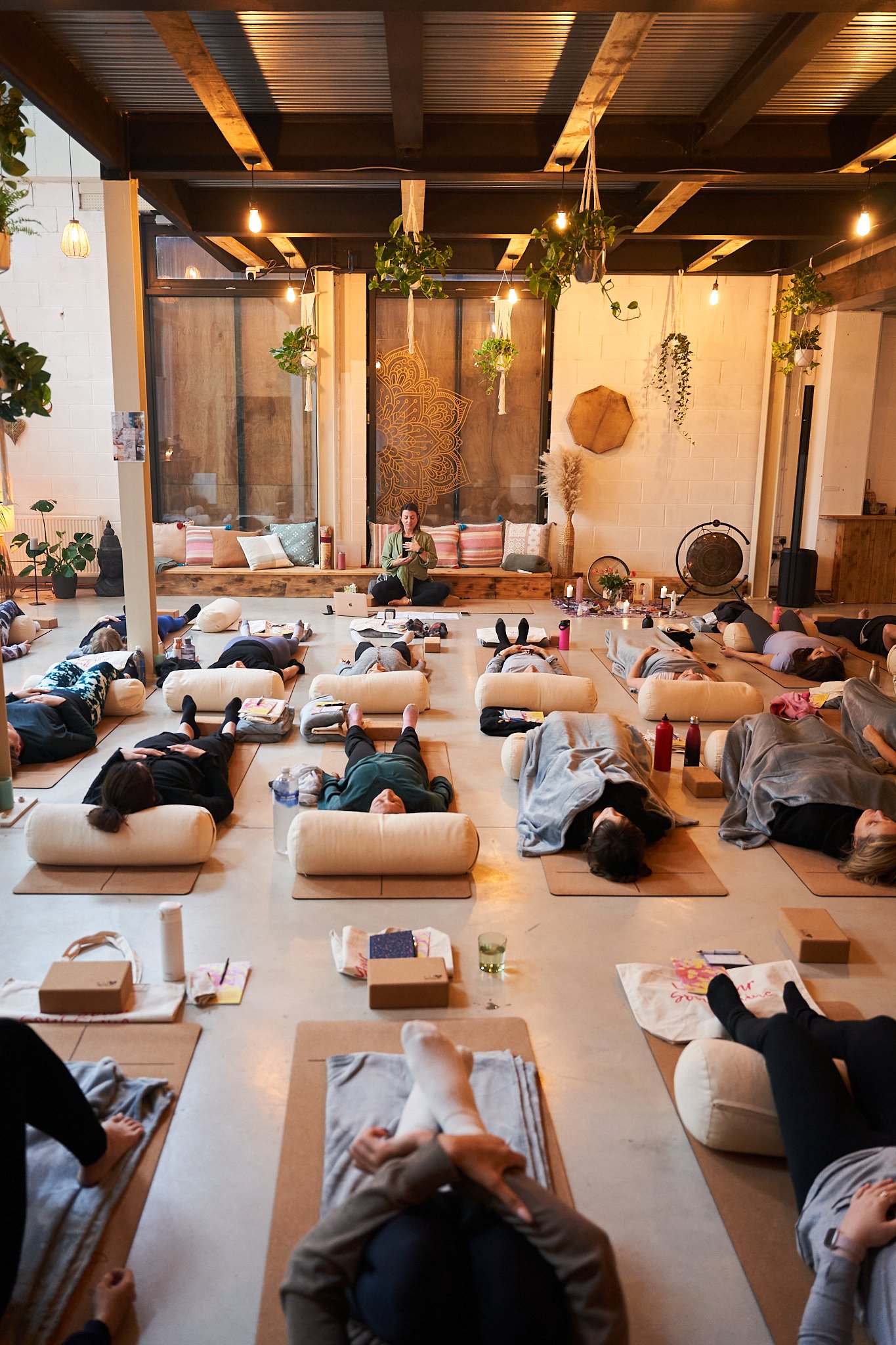 People participating in a yoga or meditation class lying on their backs on mats in a cozy, well-lit room with plants and decorative wall art.