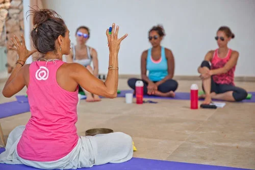 Instructor leading a meditation class with four women sitting on mats, all wearing athletic clothing and sunglasses in a bright room.