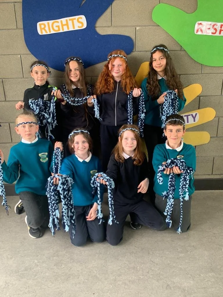 Group of young children wearing school uniforms, holding blue and white scarves, standing in front of a colorful wall display with words like 'Rights,' 'Respect,' and 'Equality'.