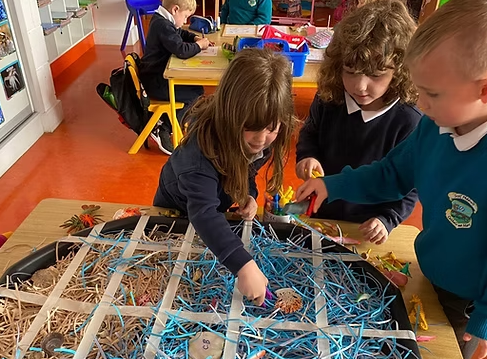 Three children gathered around a table in a classroom, examining a wire and straw model with small objects inside, in a colorful classroom setting.
