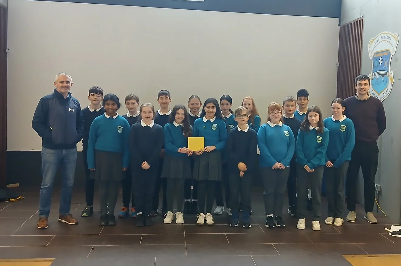 Group photo of students and two adult men in a school auditorium, with a school emblem on the wall, some students wearing blue sweaters and others in dark uniforms.