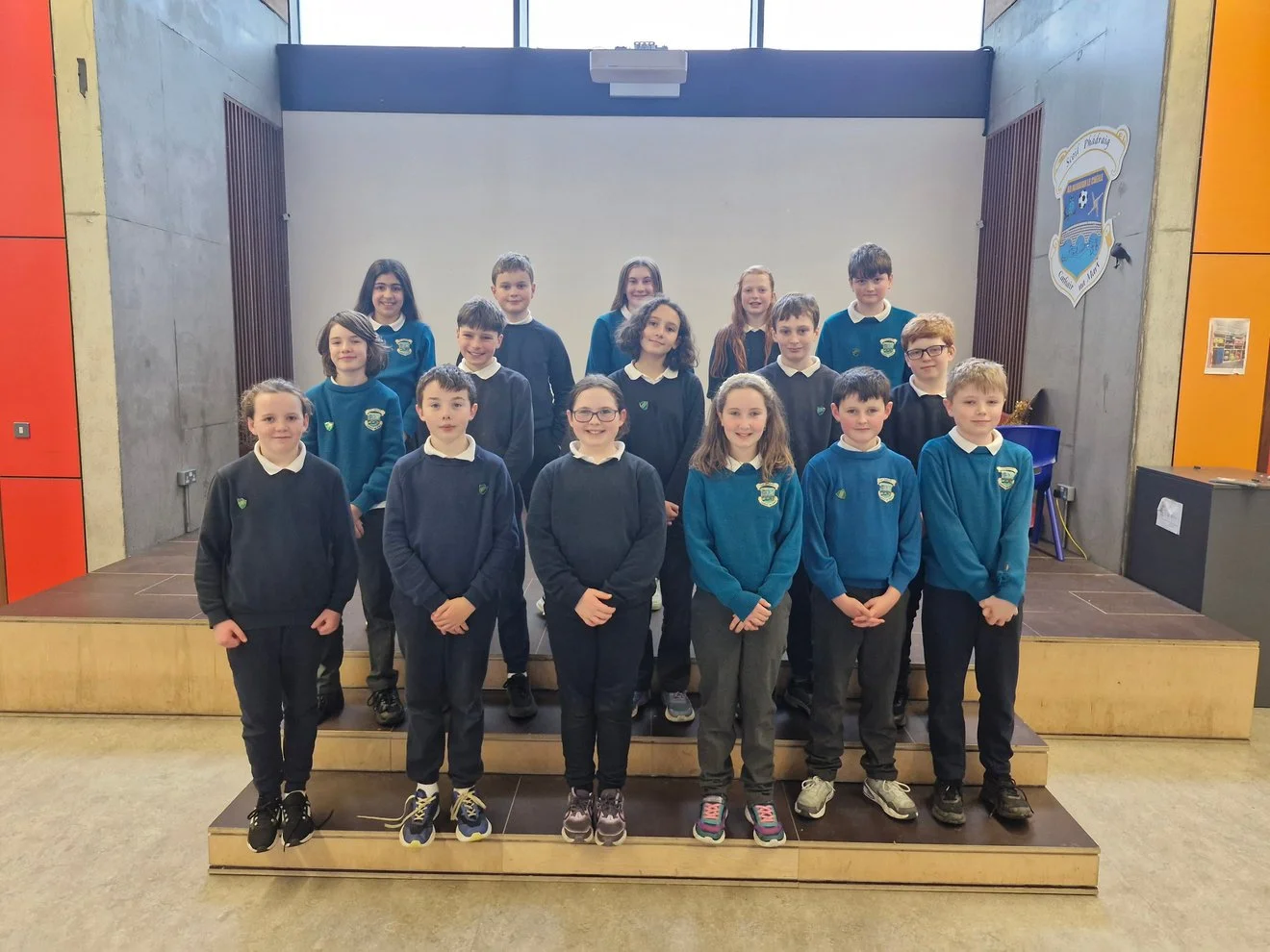 Group of 16 students standing on a stage in a school auditorium, wearing blue uniforms with school logos, posing for a class photo.
