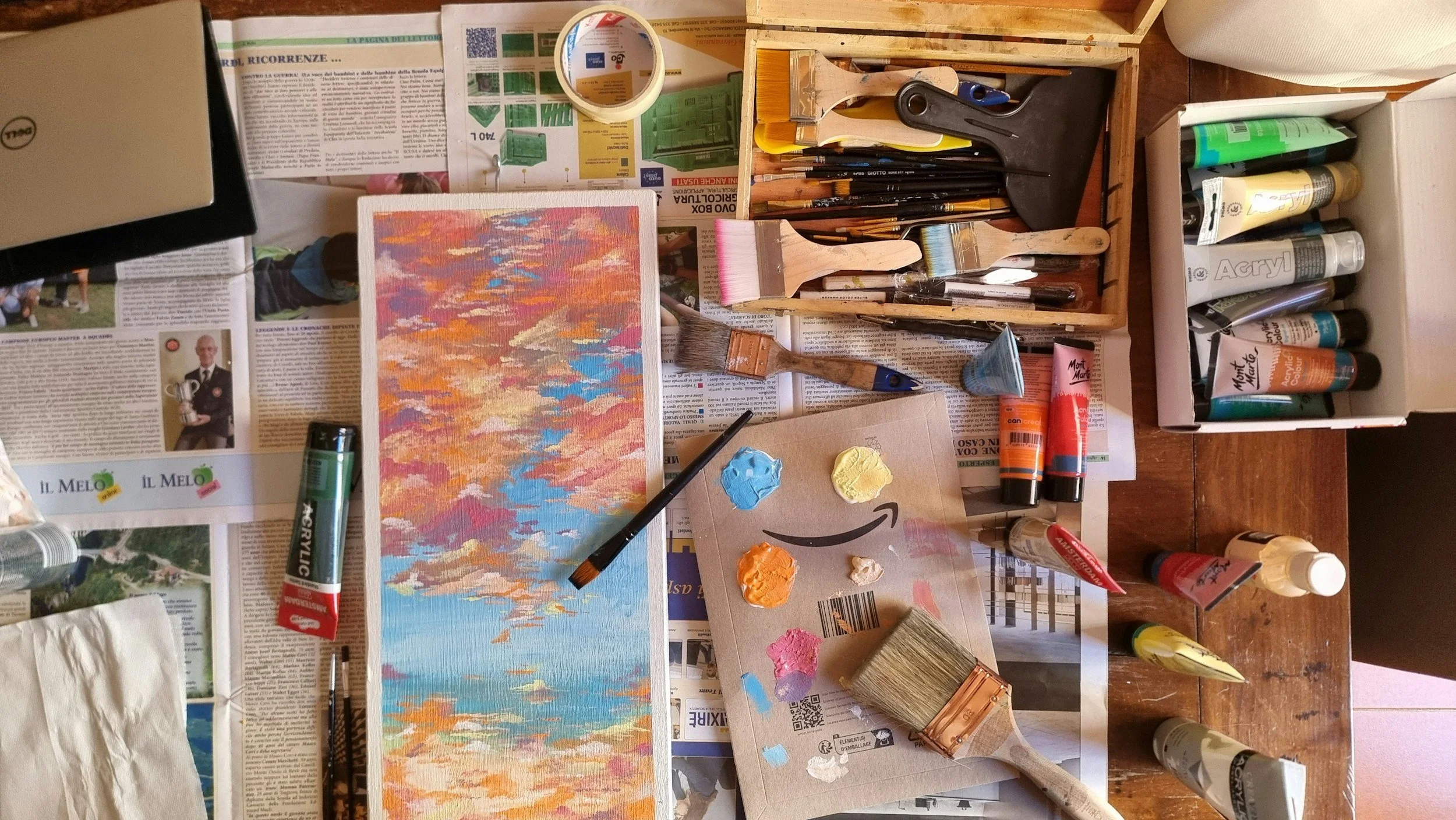 A busy artist's workspace with paints, brushes, and a partially painted landscape on a canvas. There are various art supplies, including tubes of paint, brushes, and palette knives scattered across a wooden table, with newspapers underneath.