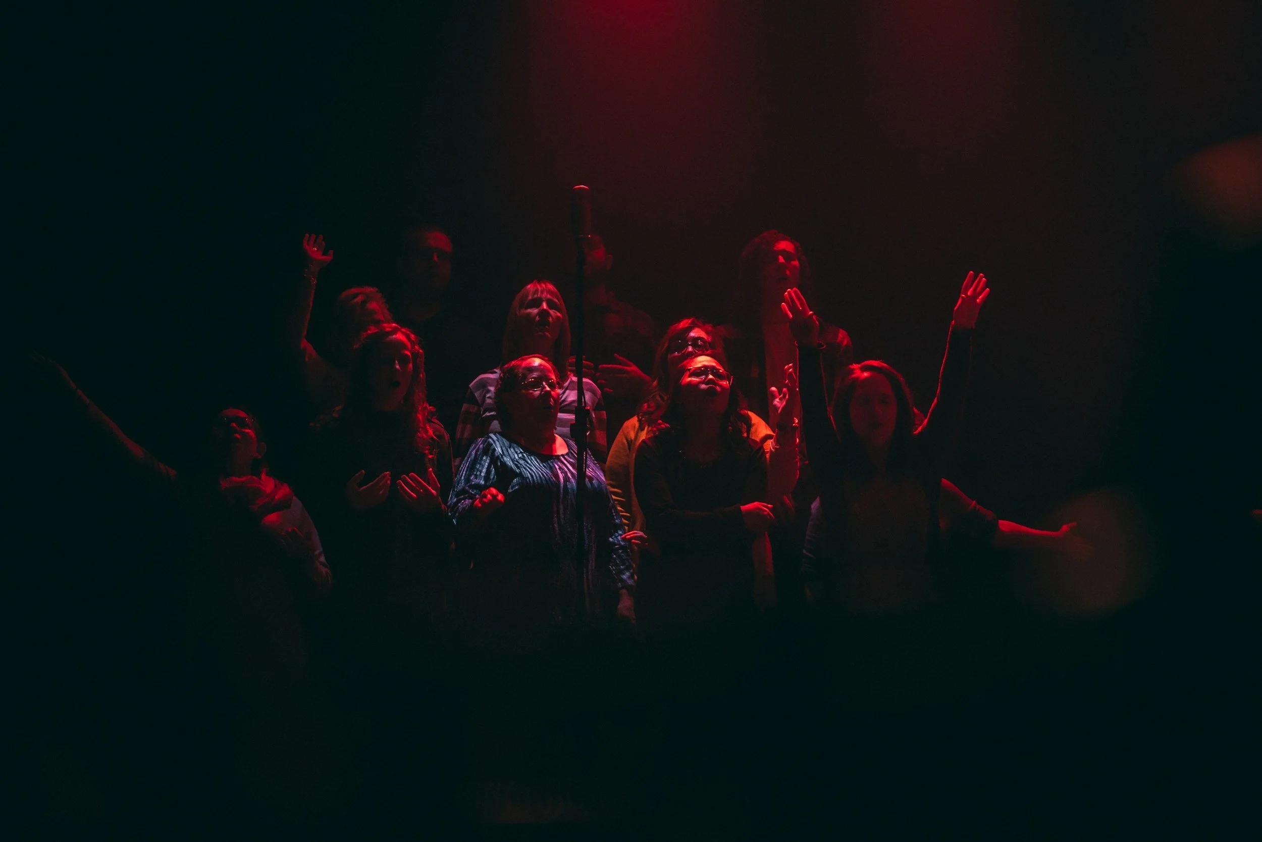 Group of people stand on stage with some raising their hands, illuminated by red lighting, against a dark background.