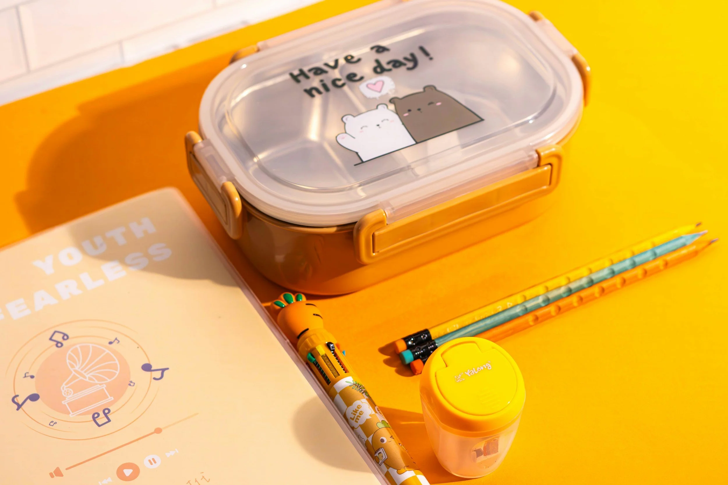 A yellow and transparent lunchbox on a yellow surface with a sticker that says "Have a nice day!" and cute bear illustrations. Surrounding the lunchbox are a yellow circle container, a yellow highlighter pen, a pencil, and a notebook with a sunflower illustration.