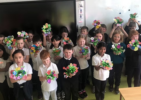 A group of young students holding colorful paper flower bouquets in a classroom.