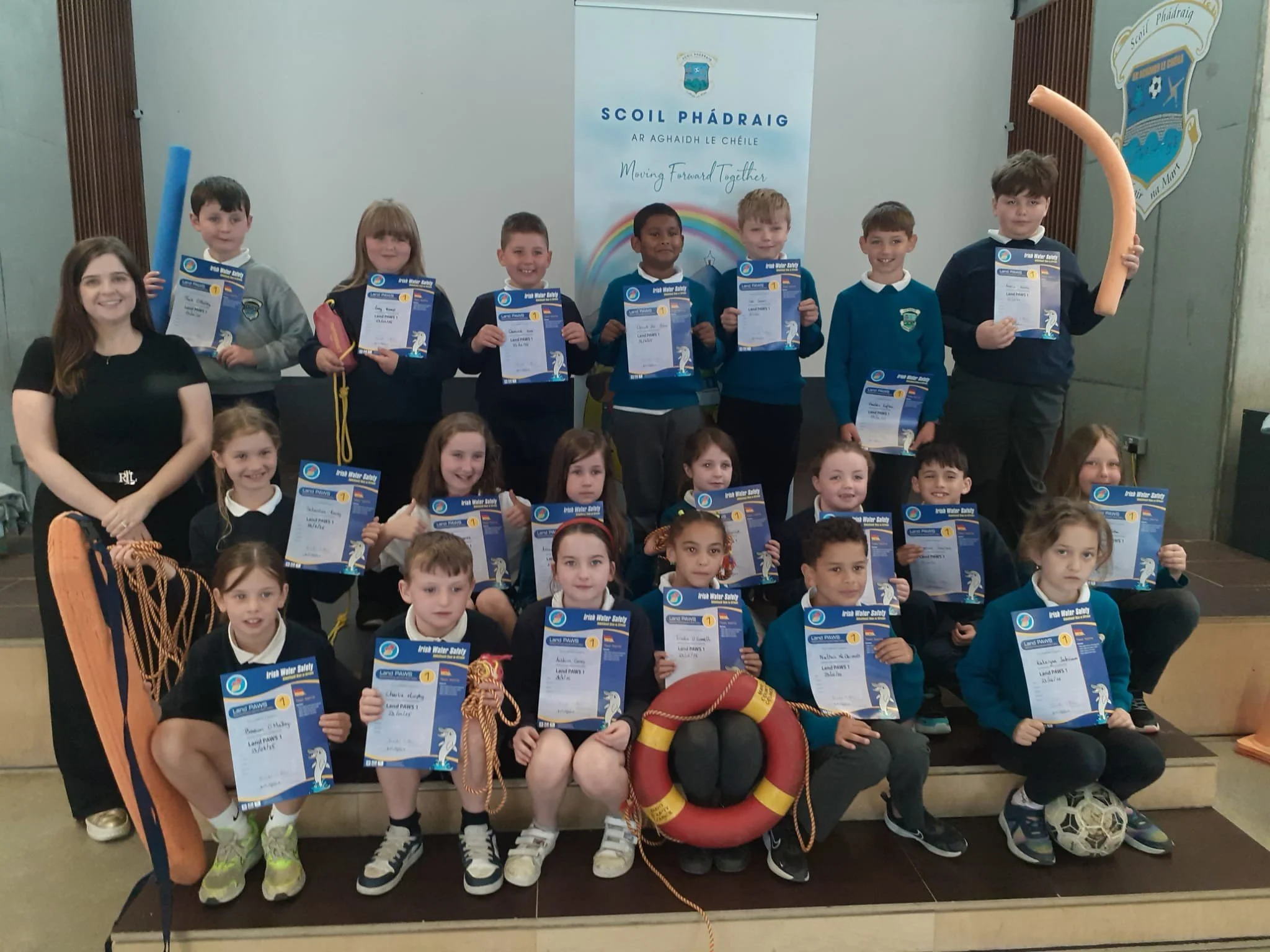 Group of children and a woman, possibly a teacher, posing with certificates and sports equipment in a school gym, with banners and a lifebuoy visible in the background.