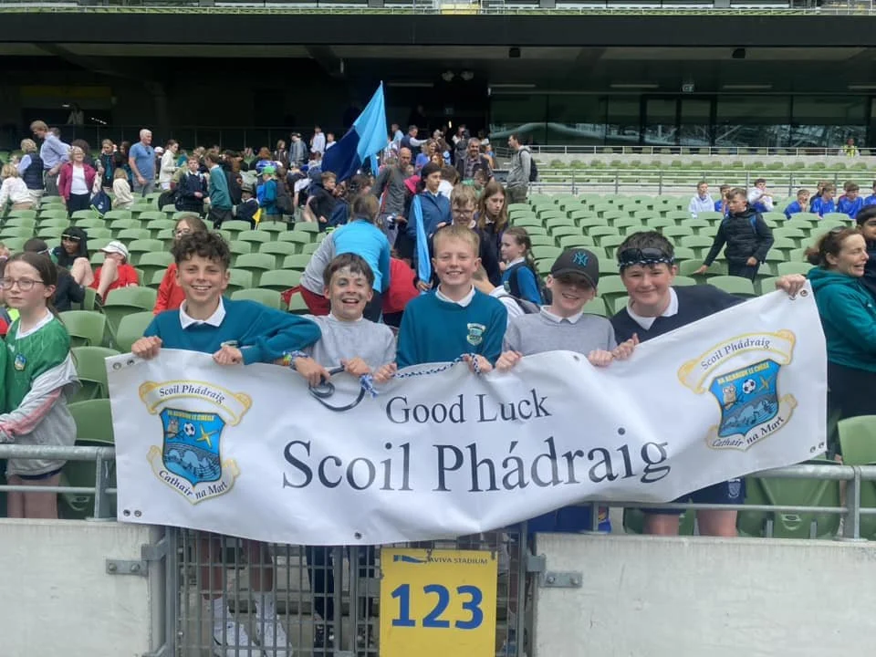 Group of children at a stadium holding a banner that reads 'Good Luck Scoil Phádraig', with other people and empty seats in the background.