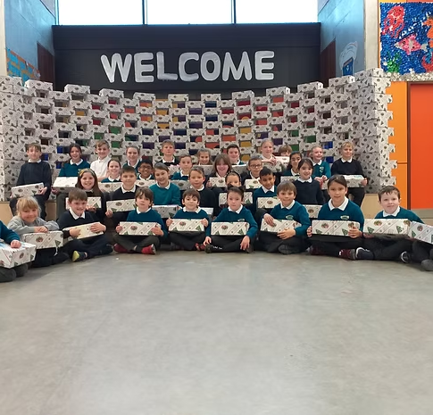 Group of young students sitting on the floor in front of a large stack of boxes or packages, with a welcome sign on the wall behind them.