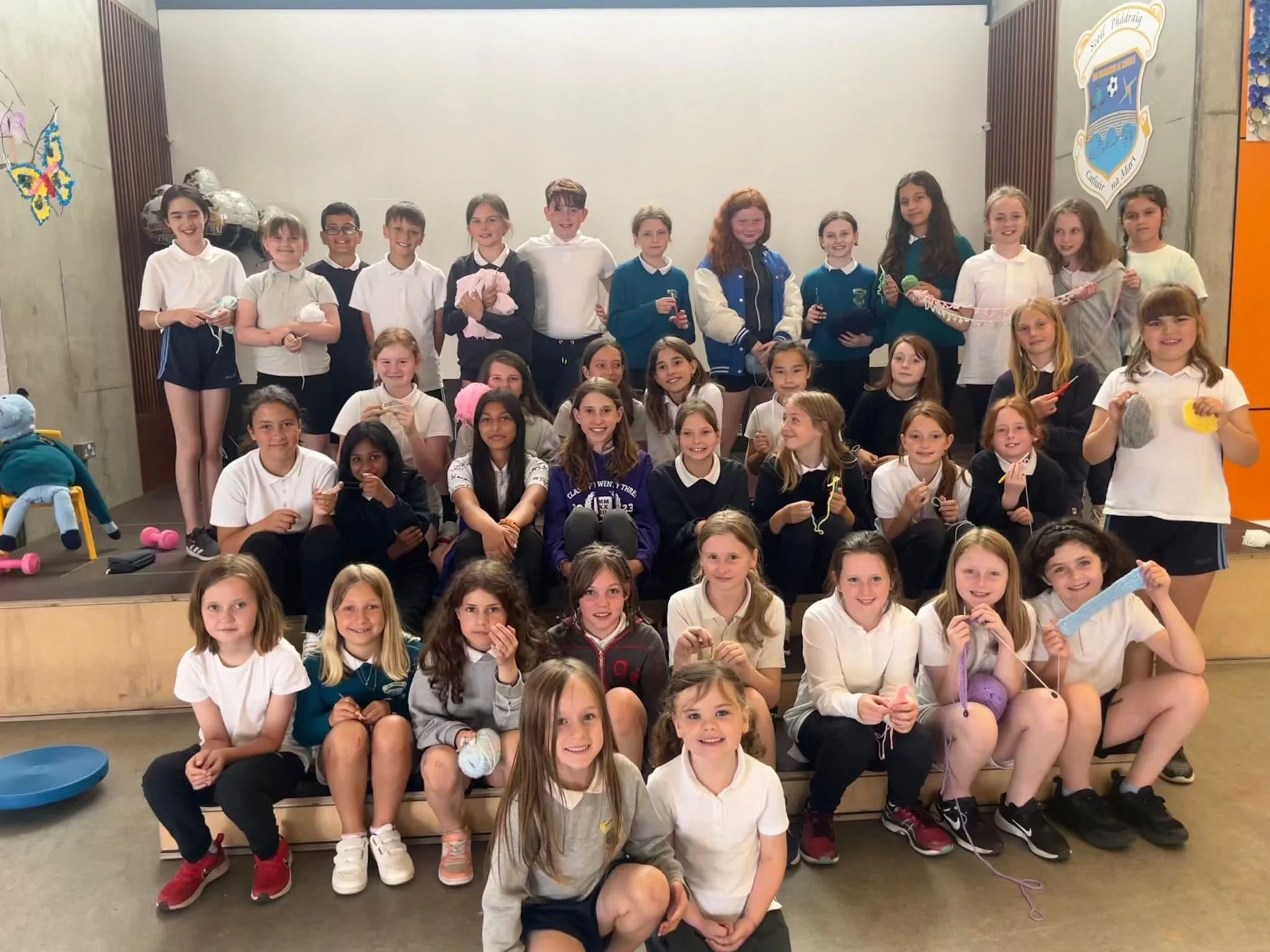 A group of diverse school children smiling, sitting and standing on a stage, some holding crafting supplies such as yarn and scissors, in a school setting.
