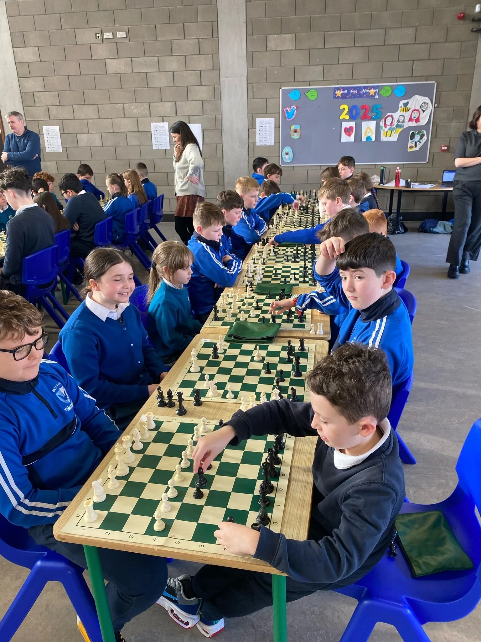 Young children playing chess in a school chess tournament, with adults supervising, decorated for 2025.