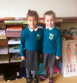 Two young girls in school uniforms standing in front of a bookshelf in a classroom.