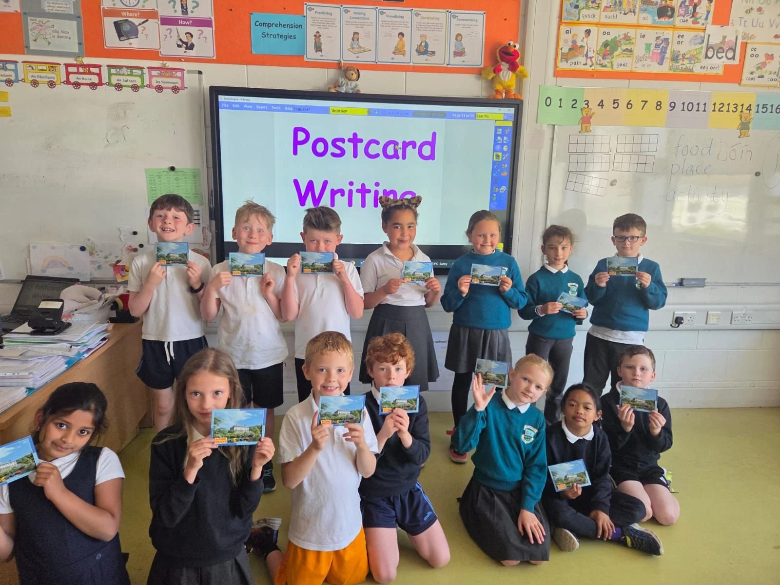 A group of elementary school students in classroom holding postcards, with a digital screen behind them displaying 'Postcard Writing'.