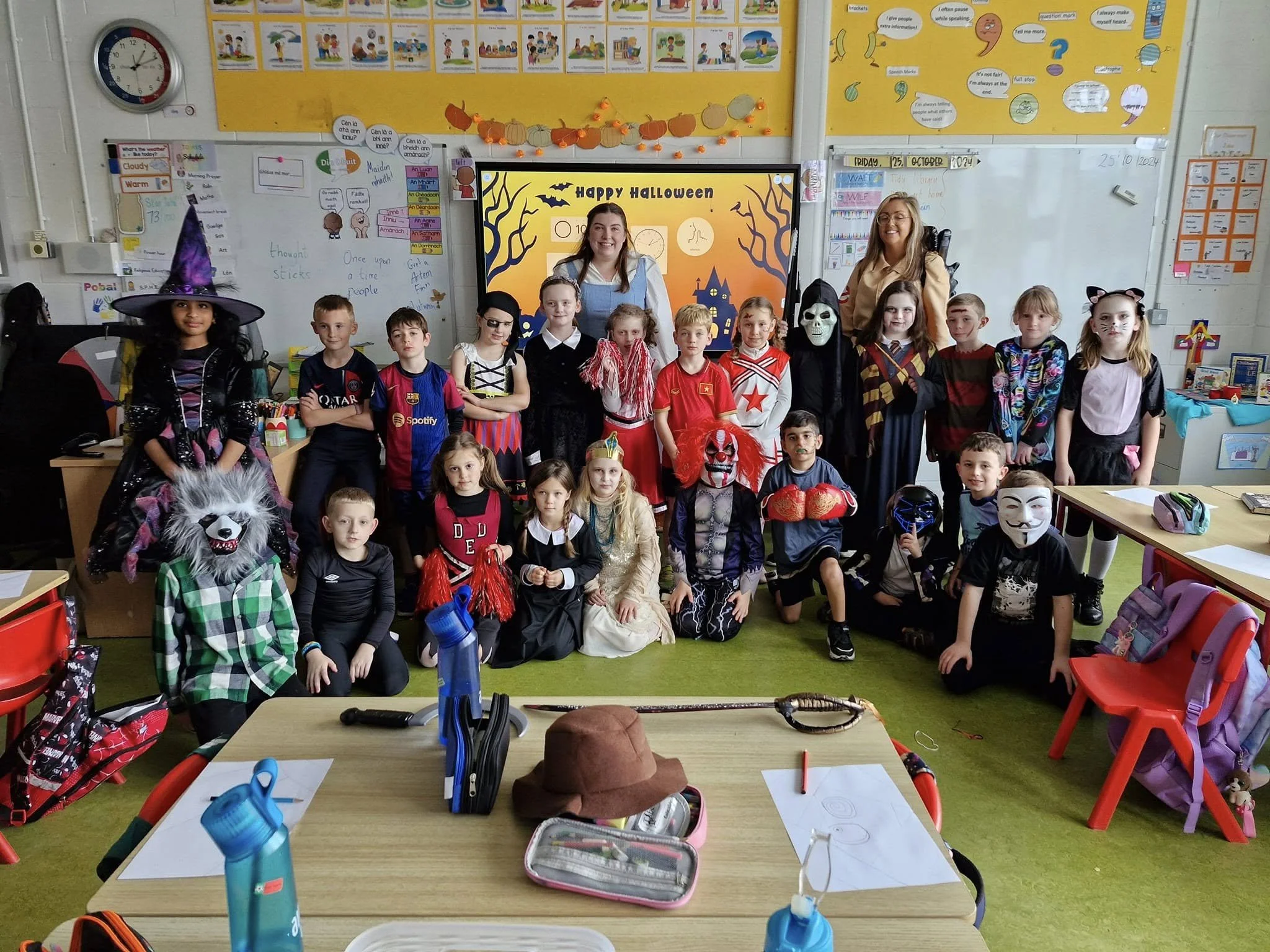 Classroom filled with children in Halloween costumes, standing in front of a yellow bulletin board and whiteboard, with classroom supplies and backpacks in the foreground.