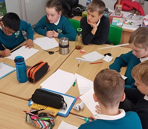 Children sitting around a table with notebooks, paper, and school supplies in a classroom.