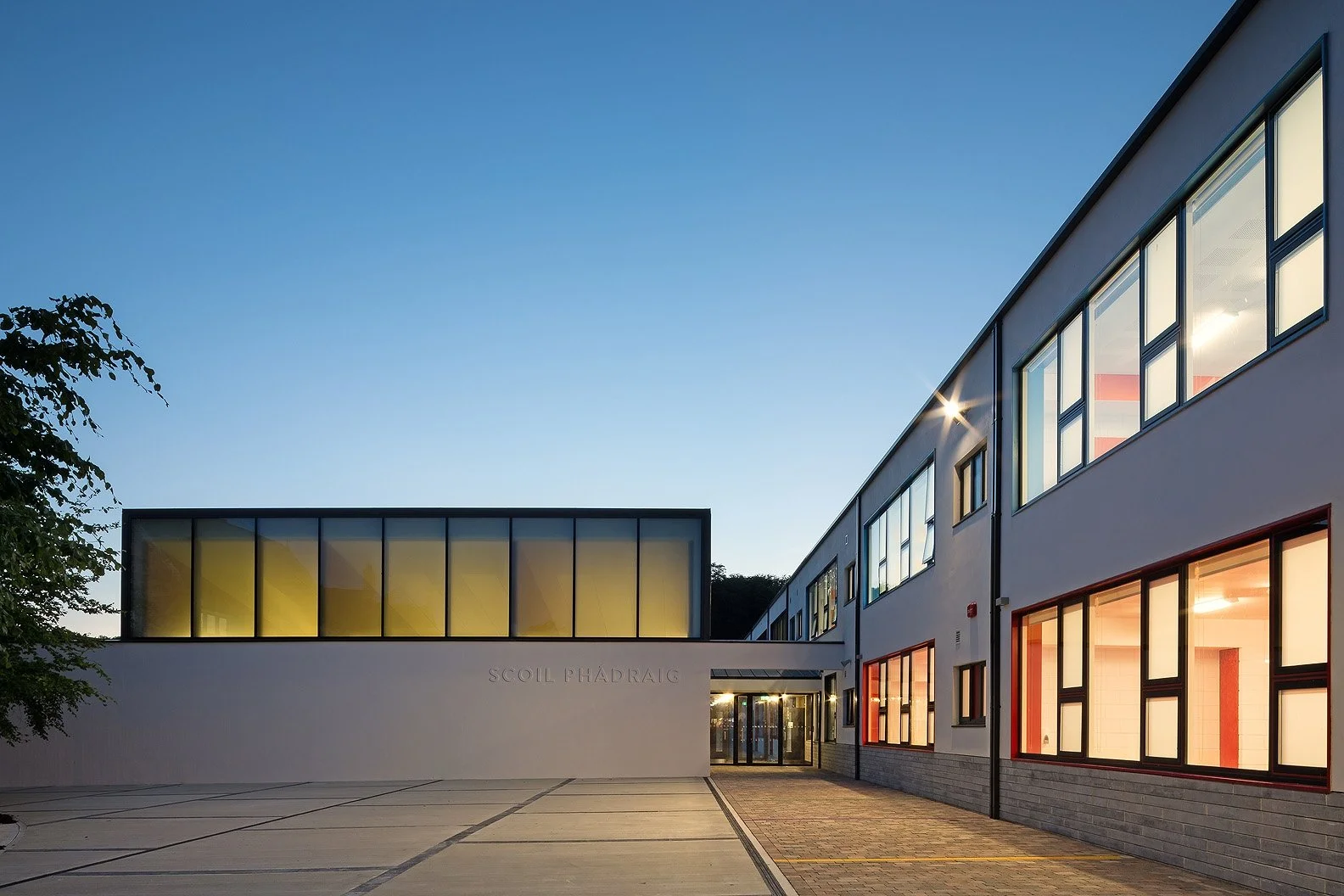 Modern educational building with large windows and a sign that reads 'SCOLL PHADRAG' on a quiet evening with a clear sky.