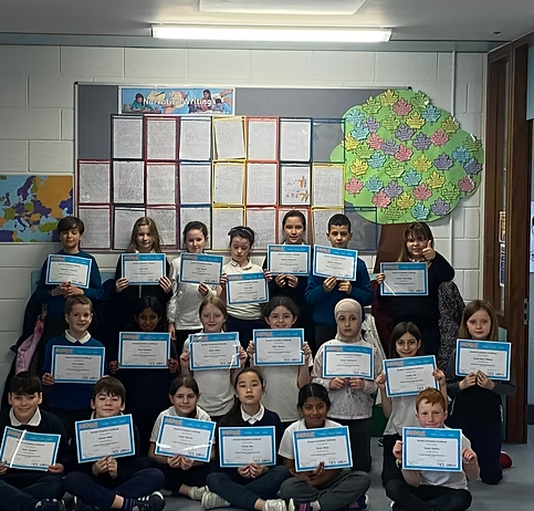 Group of young students holding certificates in a classroom, with a colorful tree and bulletin board in the background.