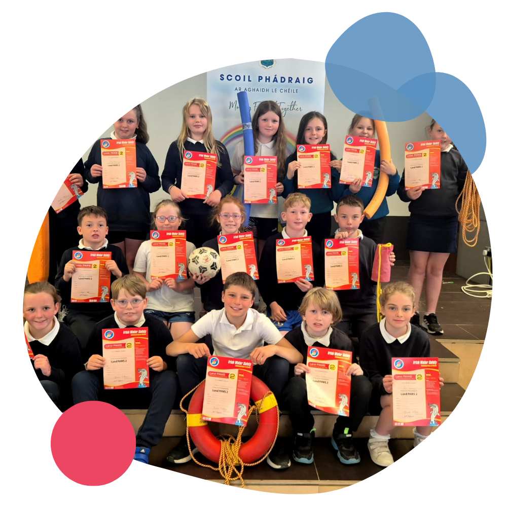 Group of children in school uniforms holding Red Cross First Aid Certificates, with some holding swim safety equipment like a lifebuoy and a soccer ball, standing on steps in a classroom or school hall.