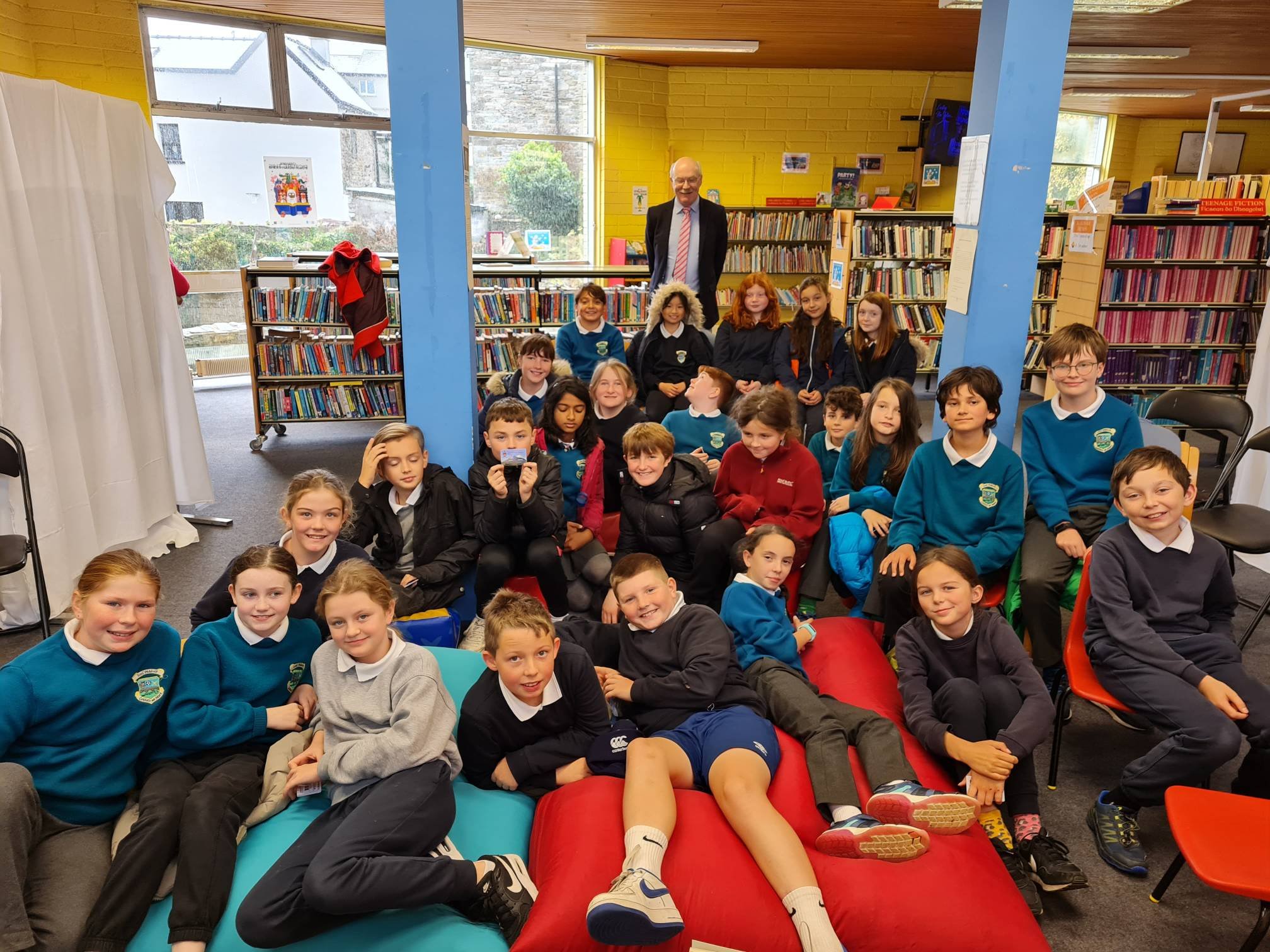 A group of elementary school children and a man standing behind them inside a library or classroom with bookshelves, large windows, and colorful furniture.