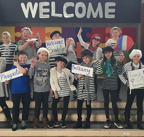 Group of young children dressed as sailors, some holding signs with welcome messages, posing on stairs in front of a colorful puzzle-piece wall and large 'WELCOME' sign.