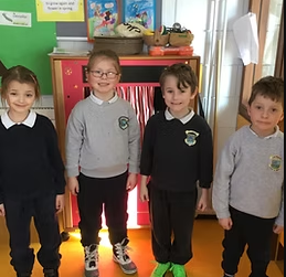 Four children standing side by side in a classroom, smiling at the camera.