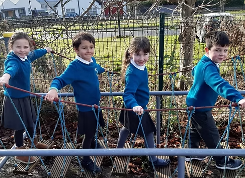 Four children wearing blue sweatshirts and dark pants are walking on a rope bridge outdoors, holding onto ropes for support, with a fence and trees in the background.
