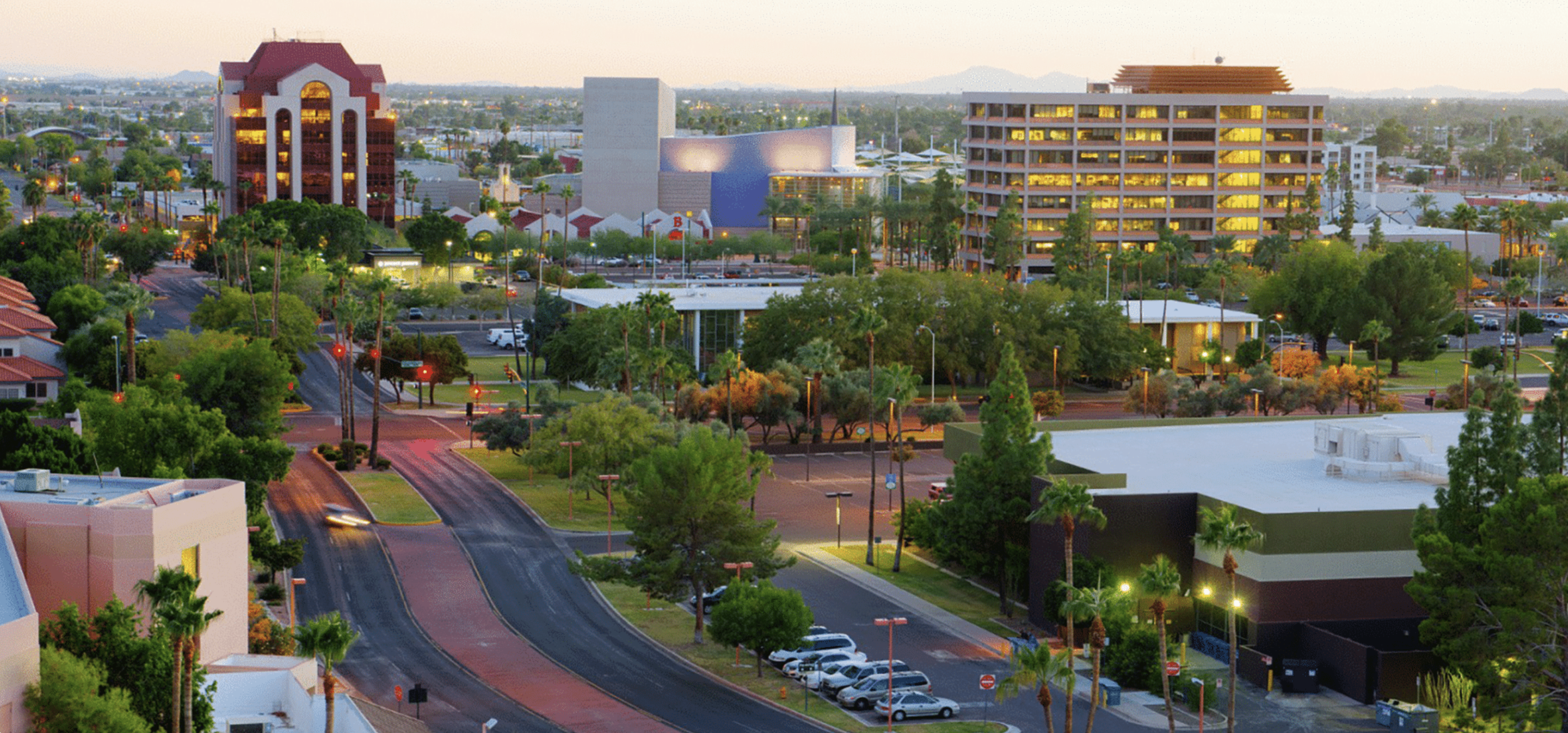 A cityscape at dusk with illuminated buildings, trees, and empty parking lots and streets.