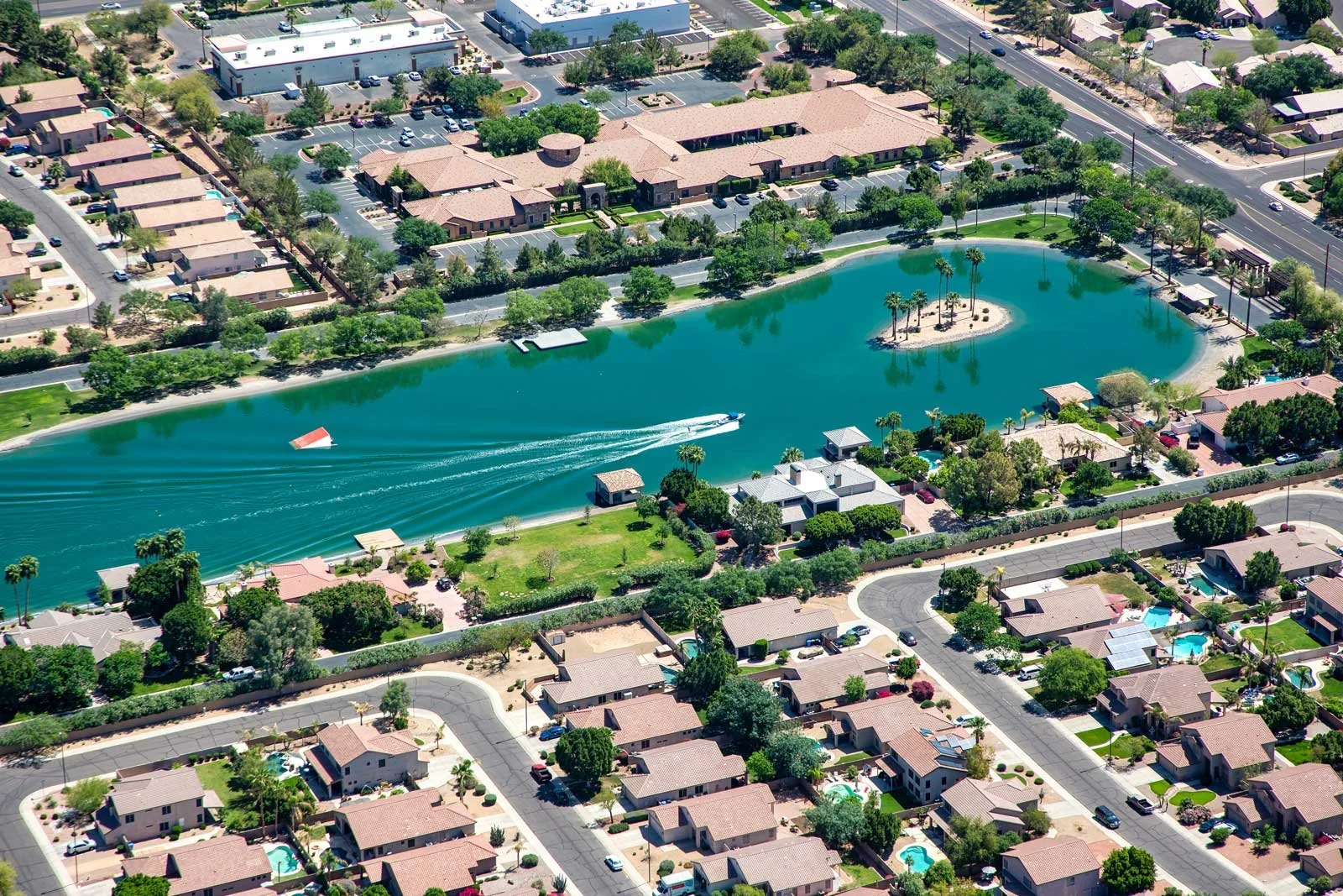 Aerial view of a residential neighborhood with a large pond, palm trees, houses with swimming pools, and surrounding streets.