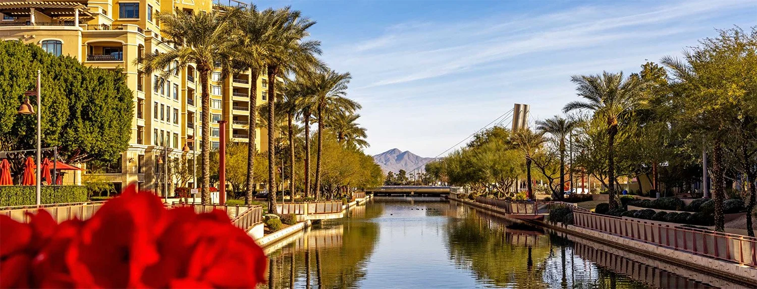 A scenic view of a canal or waterway in Scottsdale with reflections, lined by palm trees and green shrubs on both sides, with multi-story buildings on the left and a mountain in the background under a partly cloudy sky.