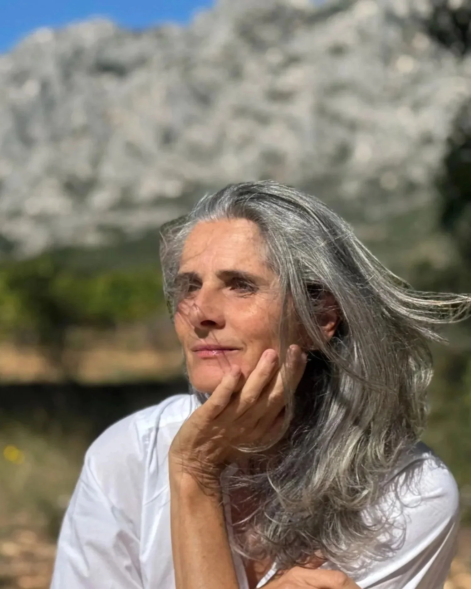 Femme aux cheveux gris, portant une chemise blanche, posant en extérieur avec un fond rocheux, regardant au loin avec une expression contemplative.