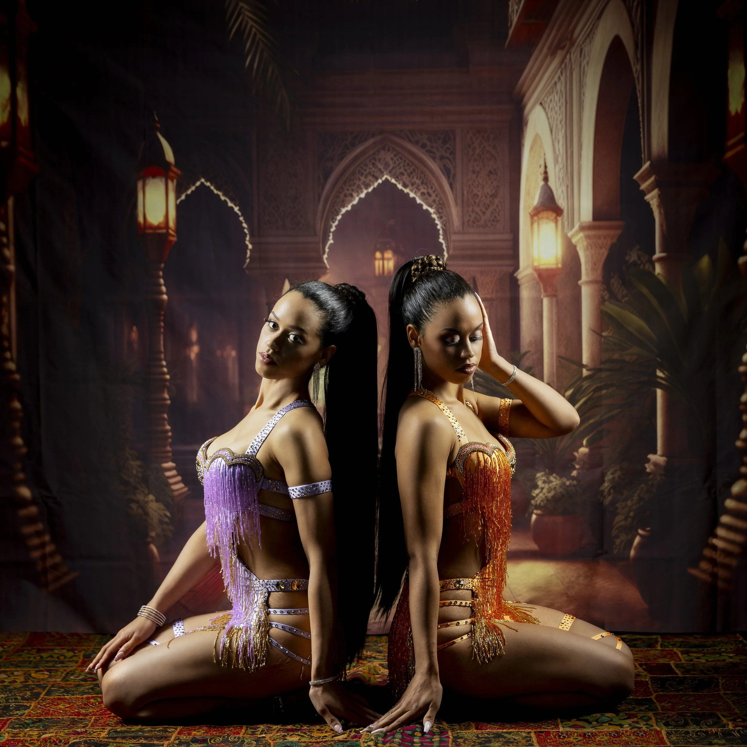 Two women dressed in colorful belly dance costumes sitting back to back on a traditional rug, in an ornate, Middle Eastern style indoor setting with arches, lanterns, and lush plants in the background.