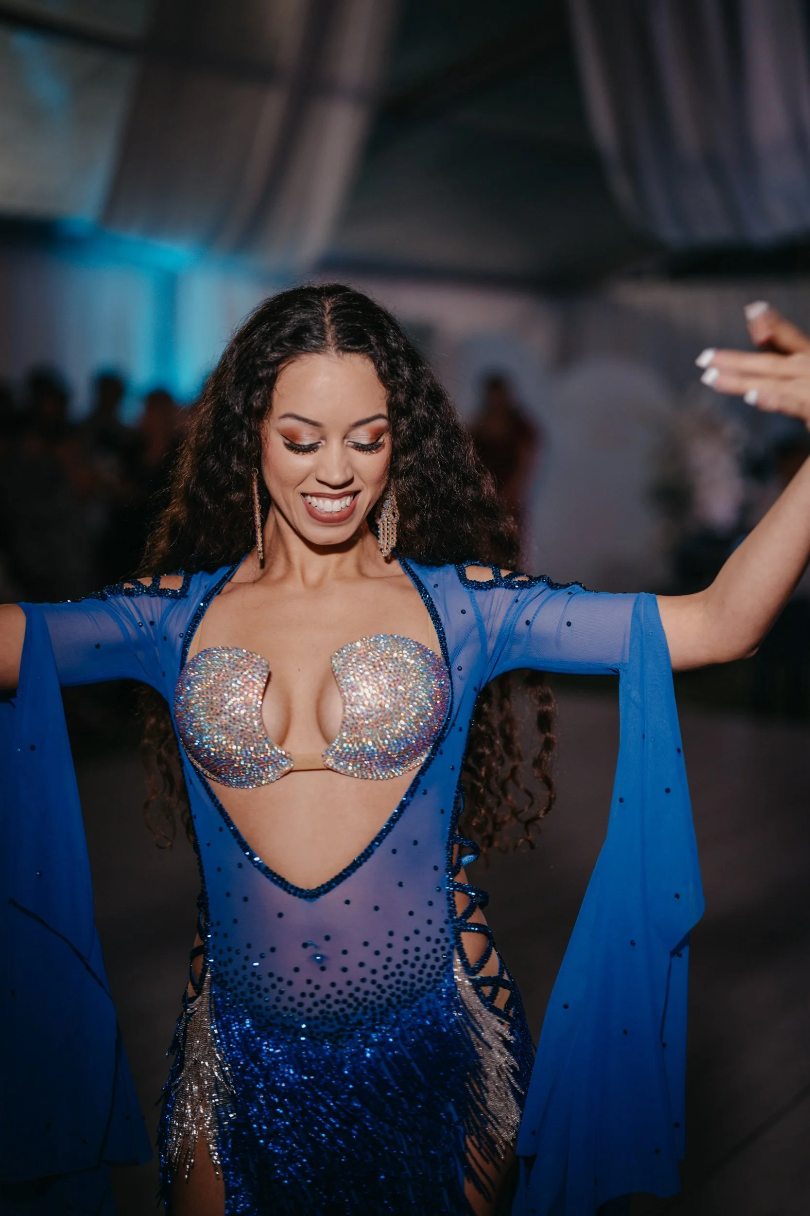 A woman wearing a blue, sparkly dance costume with a bejewelled bust, belly dancing at an indoor wedding event in Perth, smiling with her eyes closed.