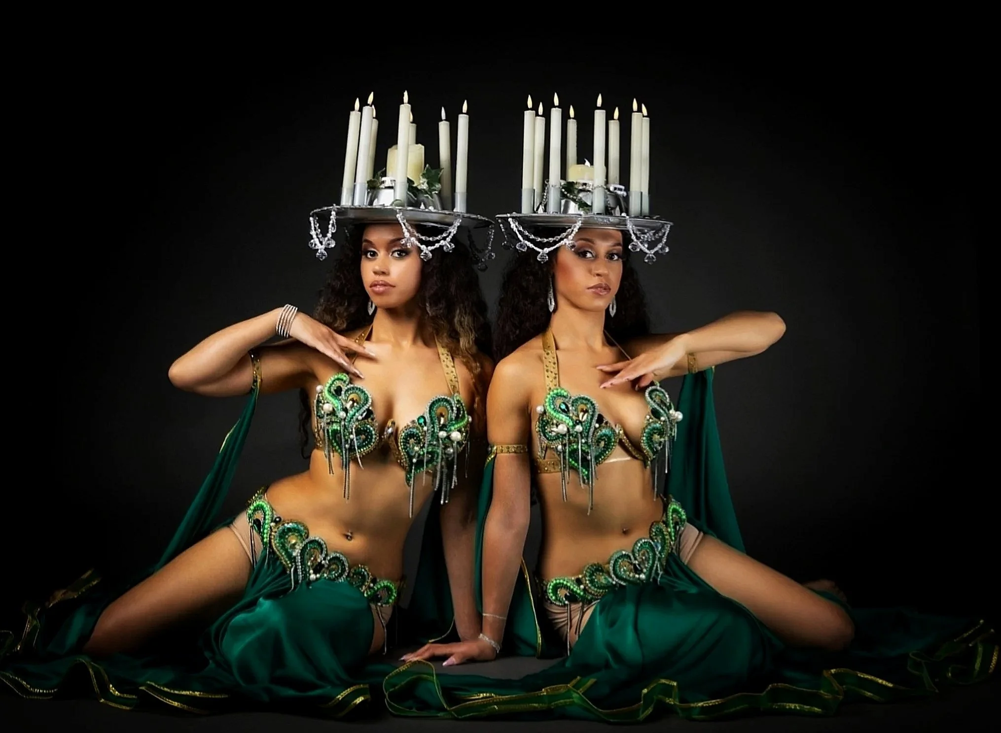 two belly dancers sitting down on the ground with emerald green belly dance costumes with sparkles, they have dance candelabra's on there head.