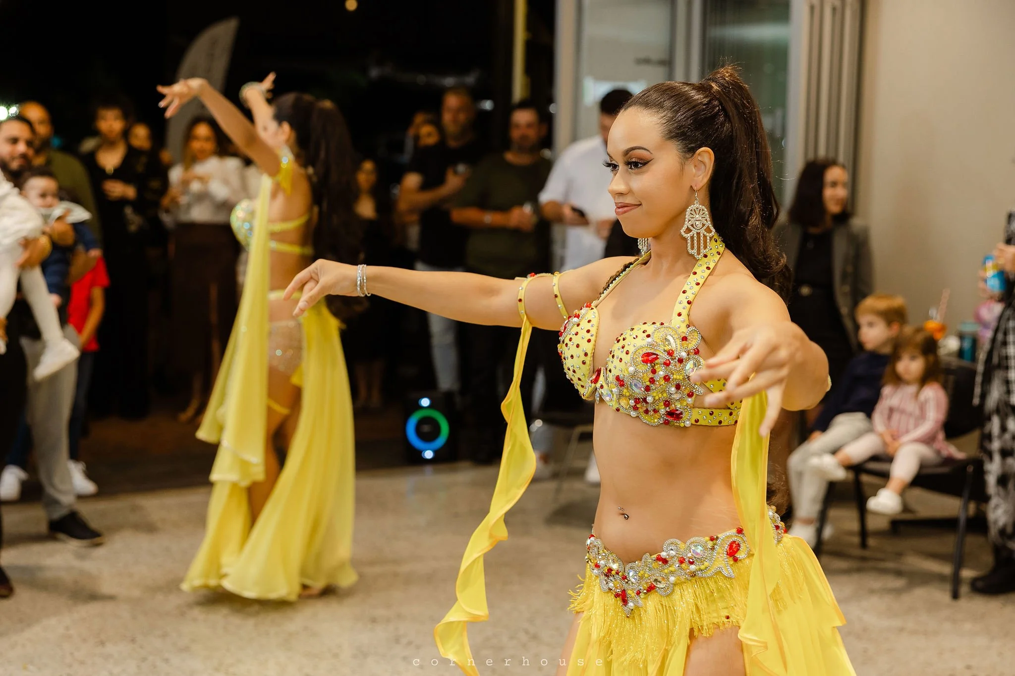 A woman in a yellow belly dance costume with jewelry performs belly dance in perth WA, for an audience at an indoor event. People are watching in the background, with some children seated and others standing.