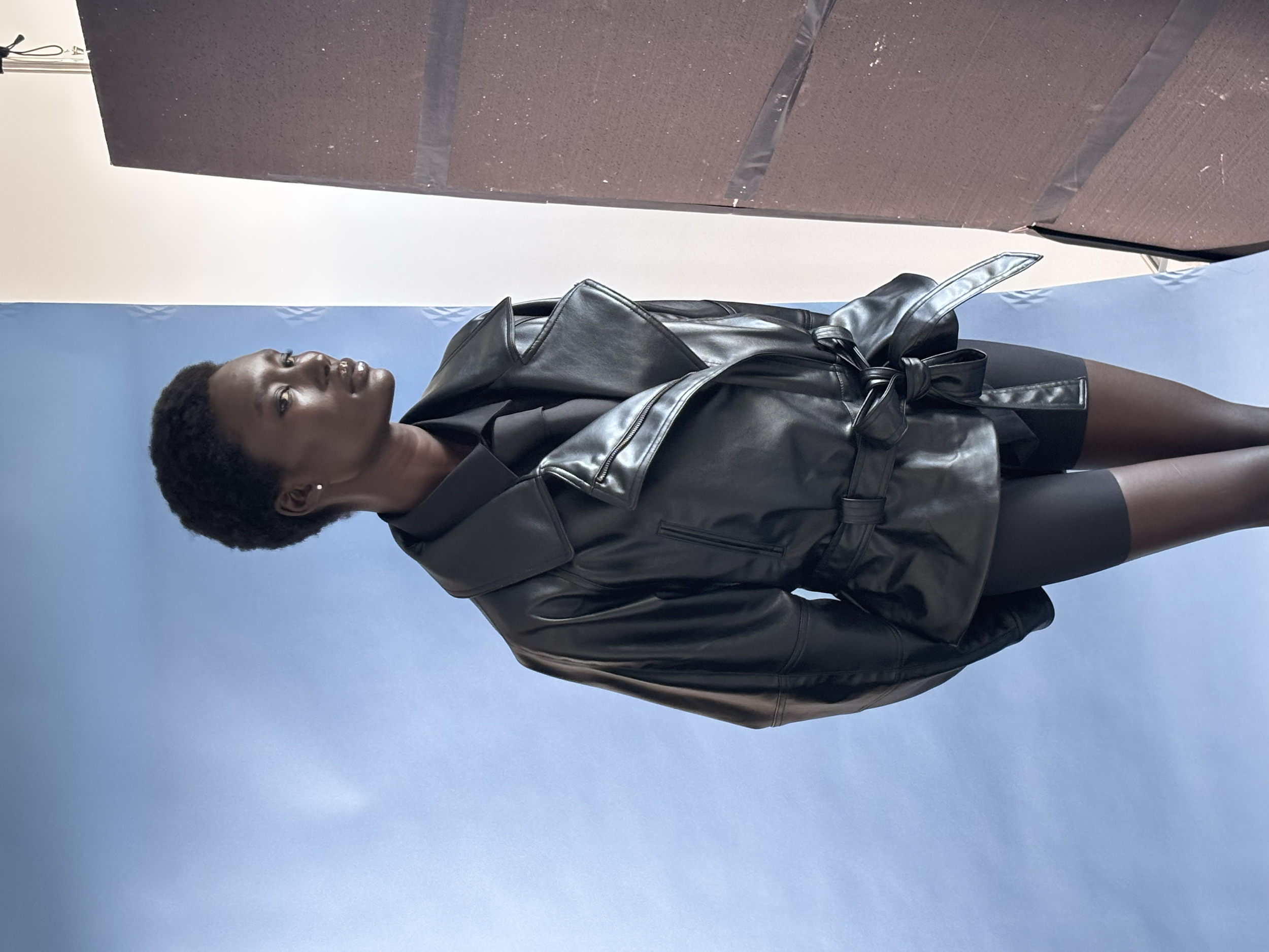 Une femme noire portant un manteau en cuir noir, une jupe noire, des collants noirs et des boucles d'oreilles.