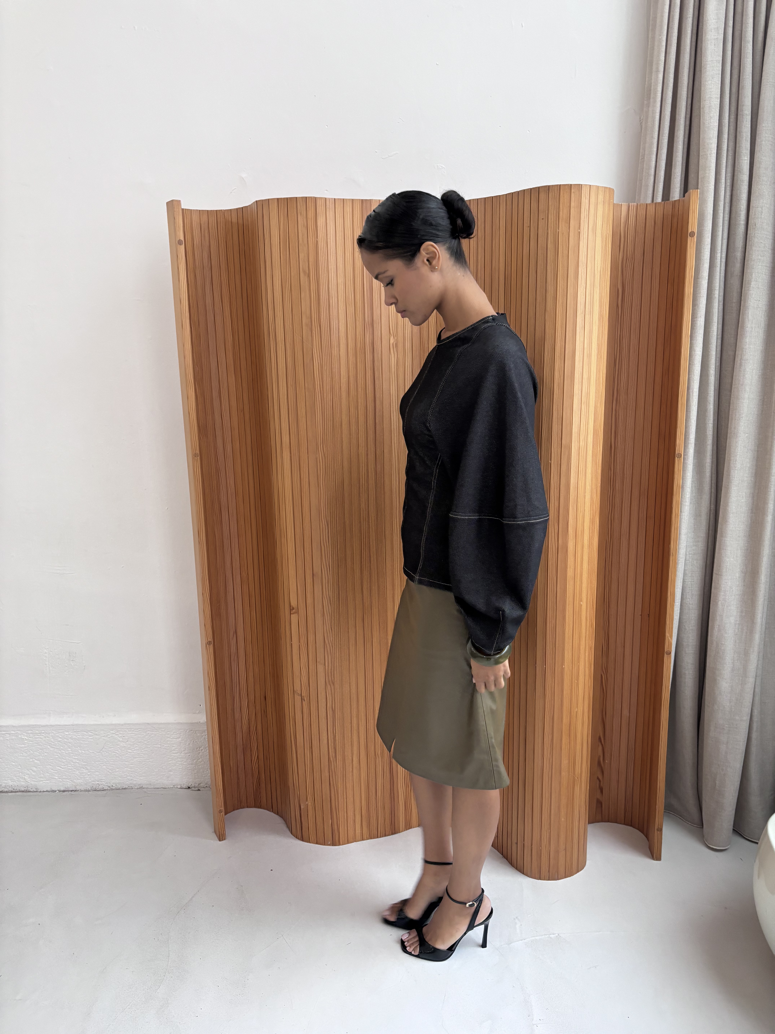 Femme debout de profil, portant une veste noire, une jupe kaki et des talons noirs, devant un paravent en bois.