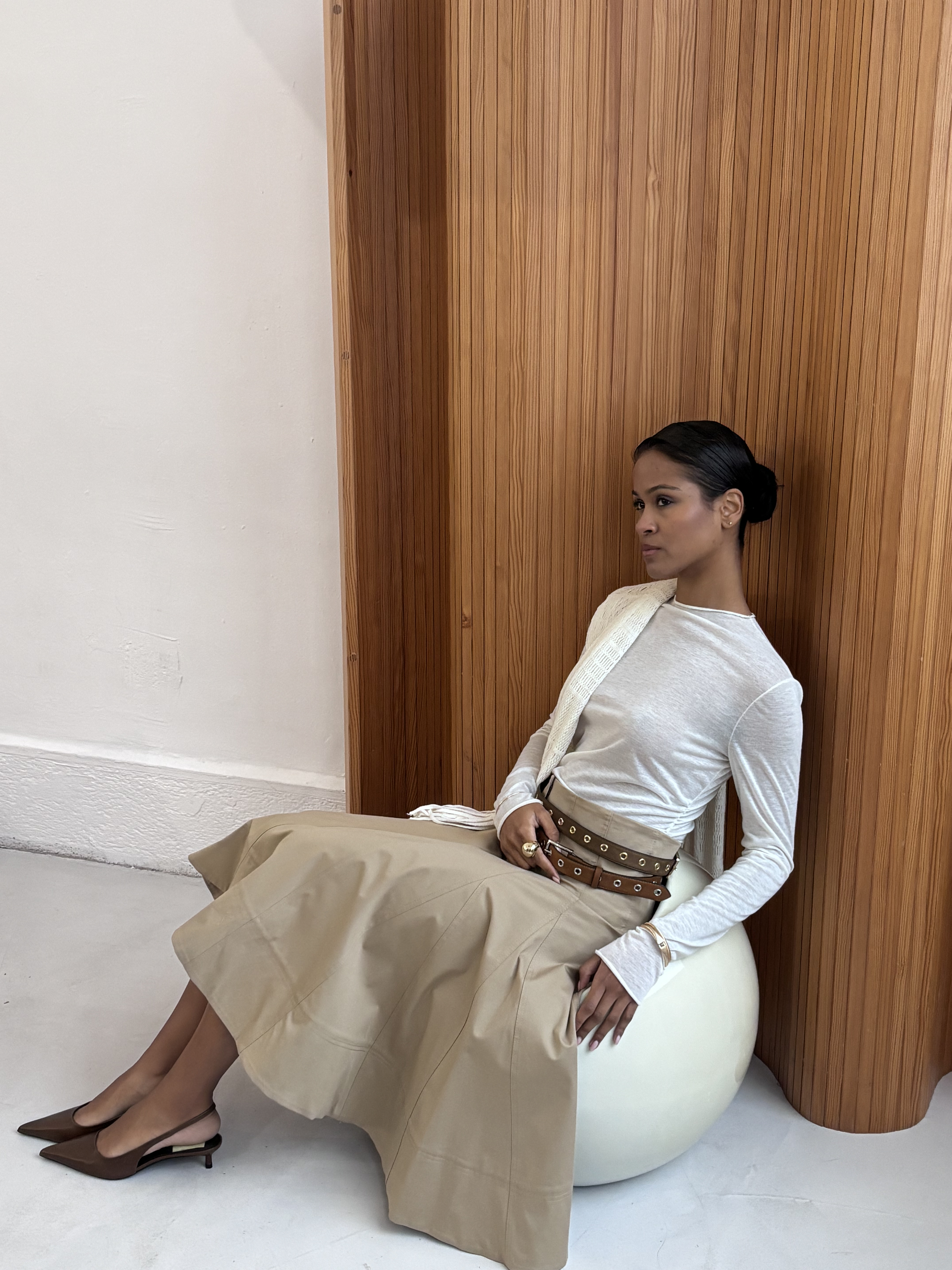 Une femme assise sur un ballon blanc, portant une jupe beige, un haut blanc, des talons marron, et avec des accessoires dorés, près d'une paroi en bois.
