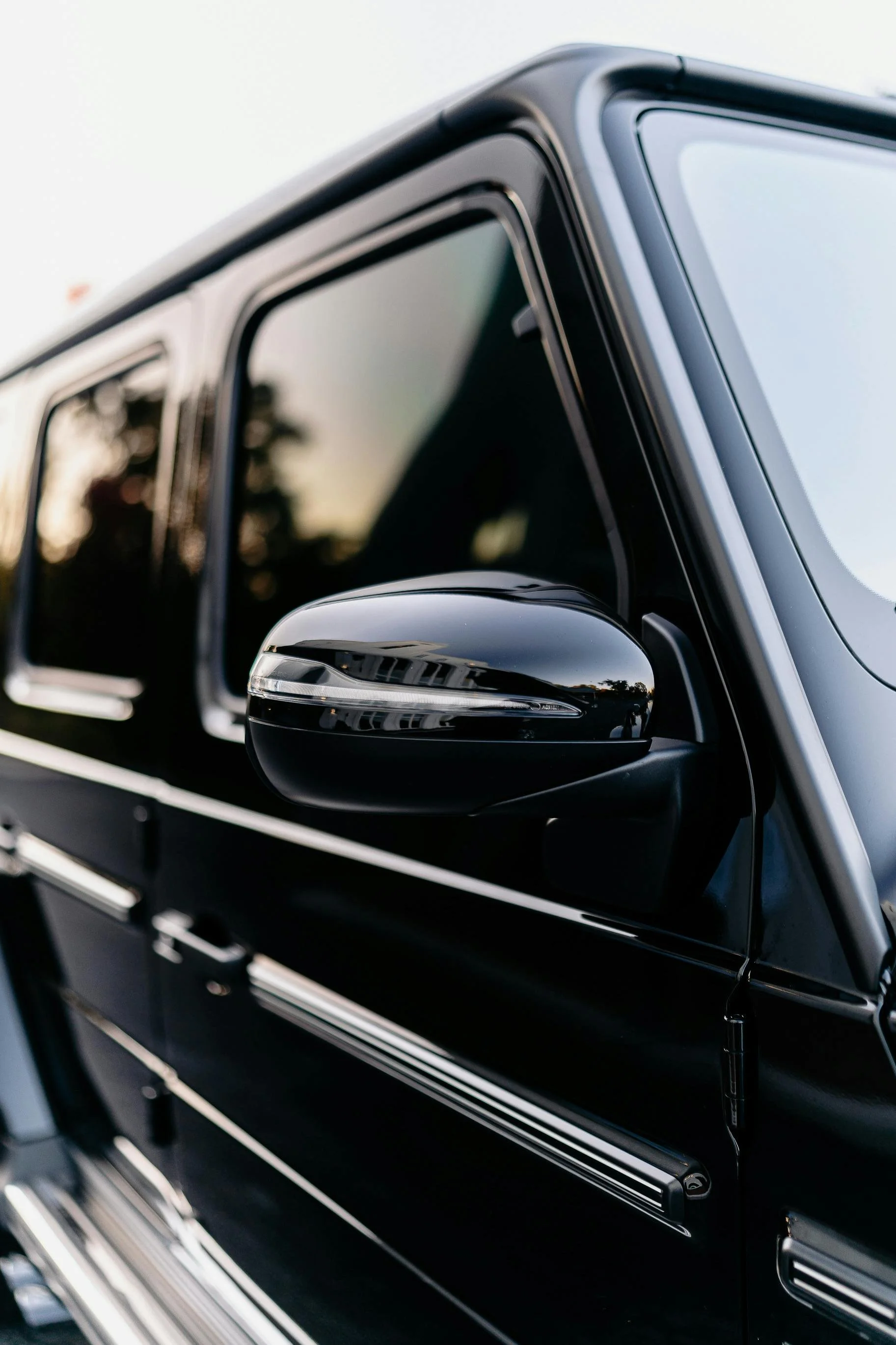 Close-up of a black luxury vehicle's side mirror and window with reflections of trees and sky.