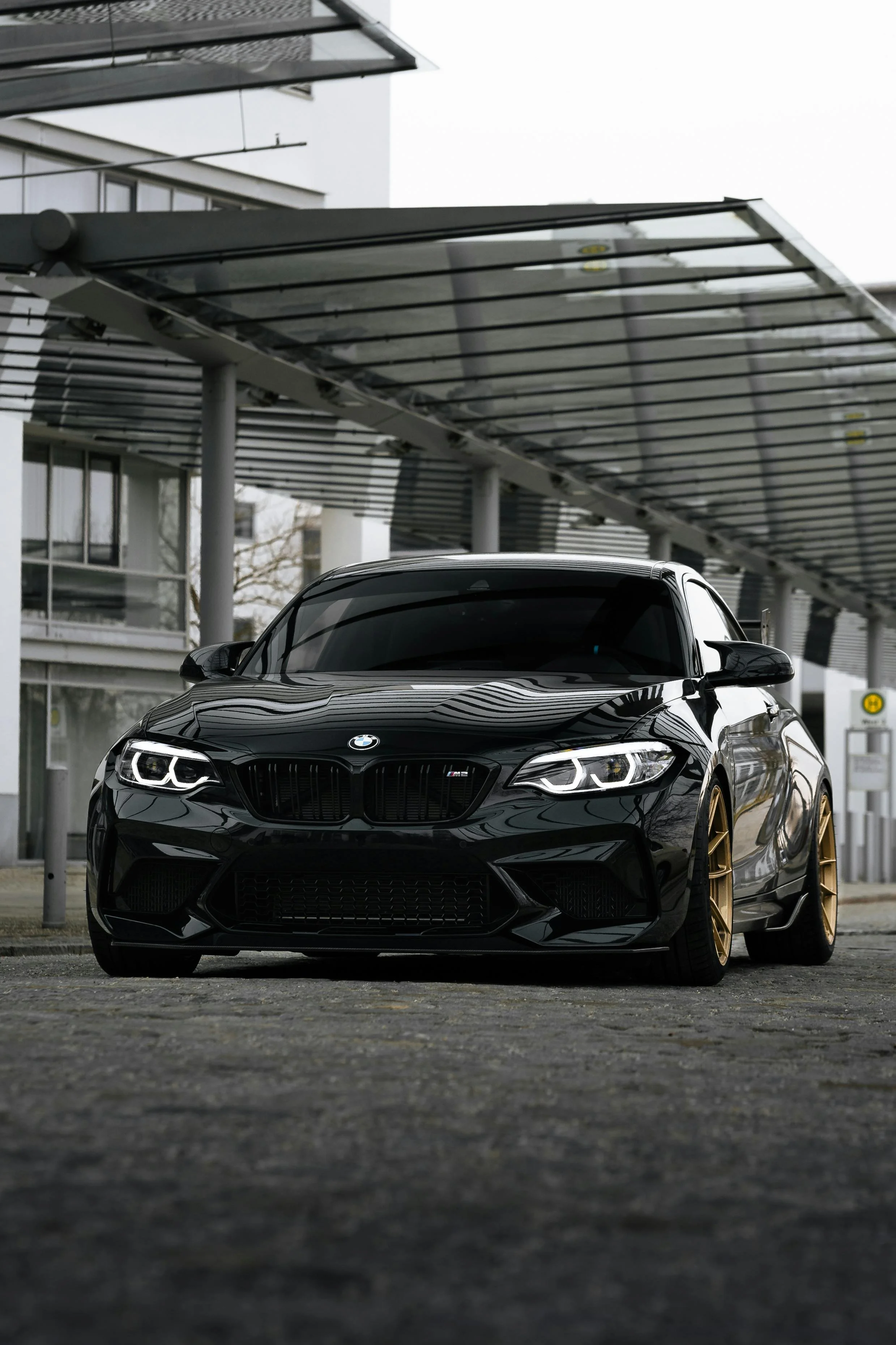 A black BMW sports car with gold wheels parked under a modern building with a glass and steel canopy.
